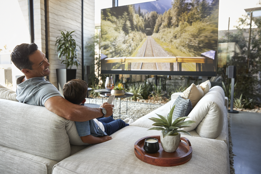 A father and son watch TV in a brightly lit living room.