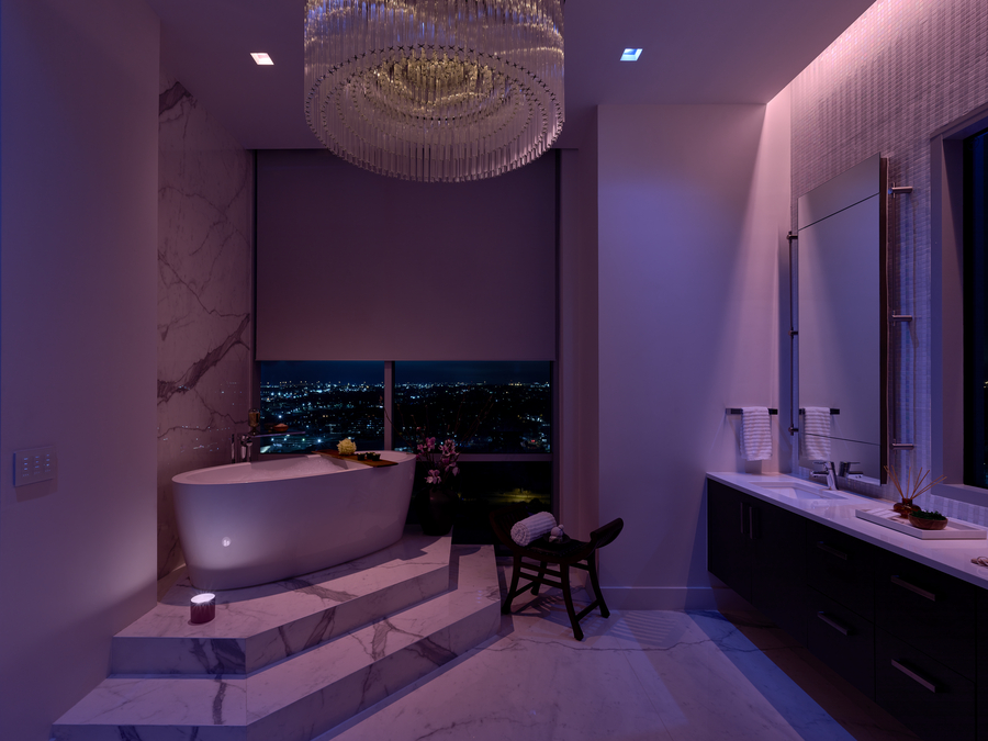 A bathroom awash in Lutron Ketra’s soft violet lighting with Lutron shades partially lowered next to a raised bathtub.