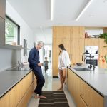 A couple stands in the middle of their kitchen wearing pajamas while their young child runs towards them. The kitchen is illuminated by Savant lighting.