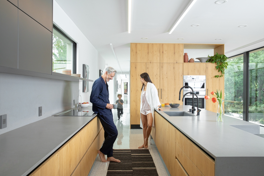 A couple stands in the middle of their kitchen wearing pajamas while their young child runs towards them. The kitchen is illuminated by Savant lighting.