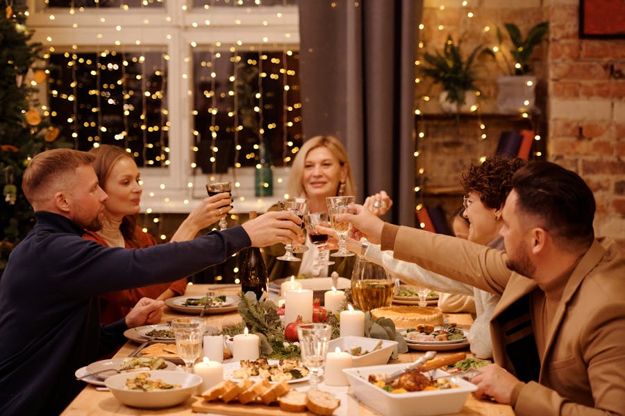 A group of people raising glasses in a festive toast around a candlelit dinner table with holiday decorations and warm string lights.